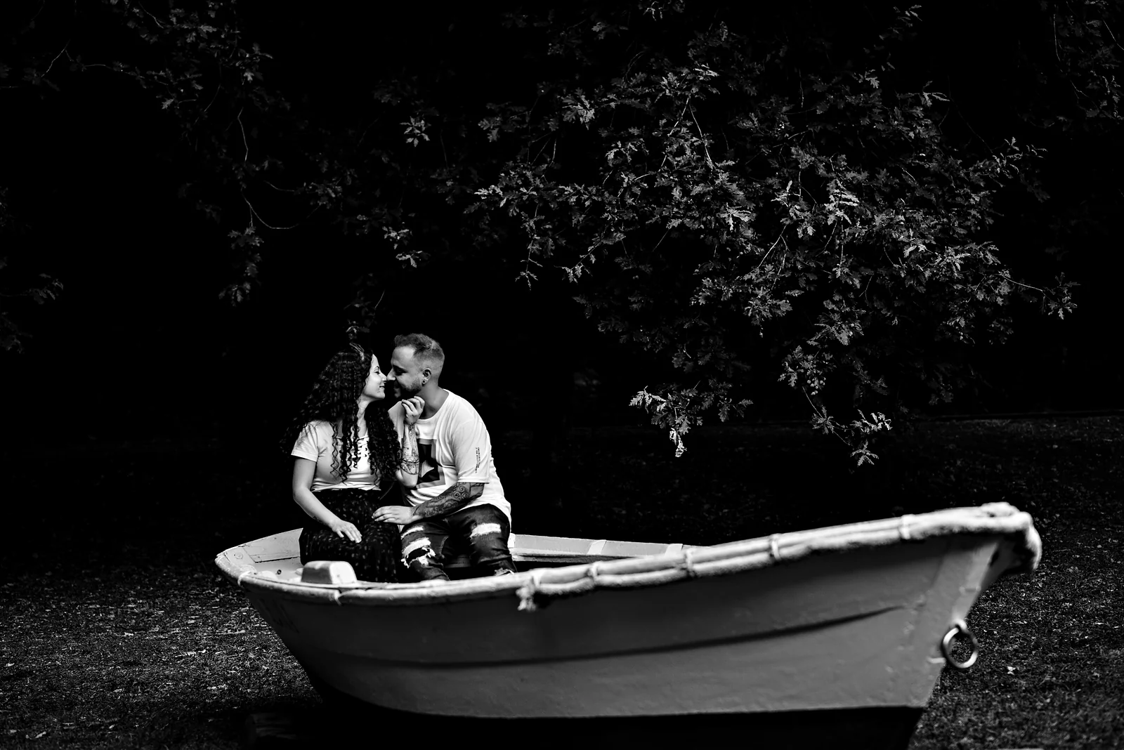 Pareja sentada dentro de una barca varada en la hierba, de noche, sobre un fondo negro.