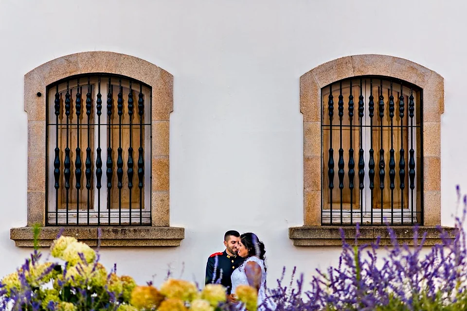 Novios besándose ante una fachada blanca con dos ventanas de granito, entre flores amarillas y lavanda.