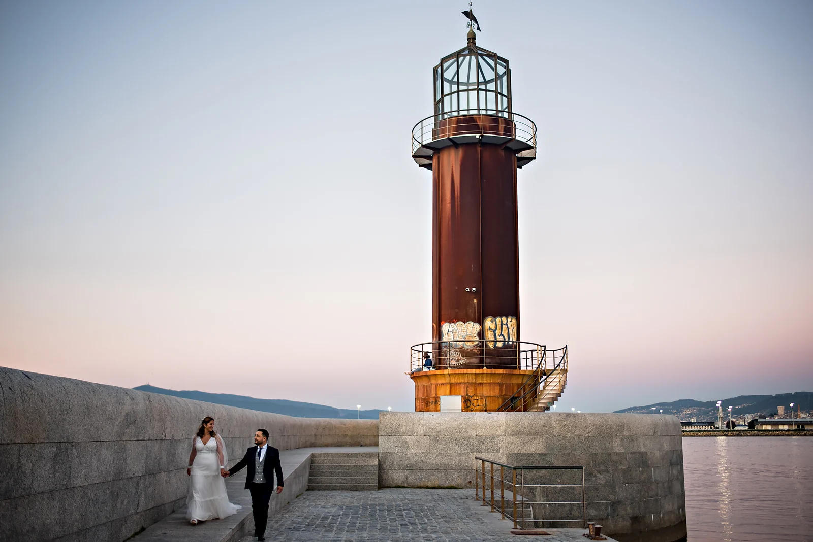 Novios caminando por un dique de piedra junto a un faro oxidado, con el cielo rosa del atardecer.