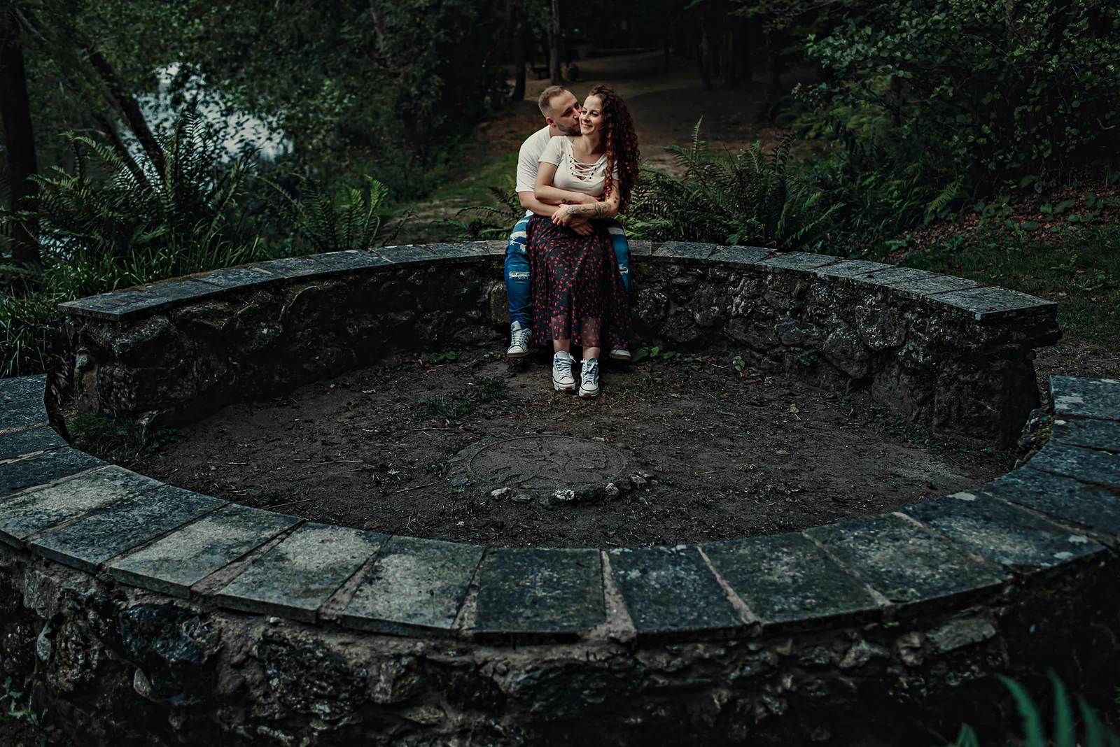 Pareja sentada en el borde de una fuente circular de piedra, rodeada de bosque.