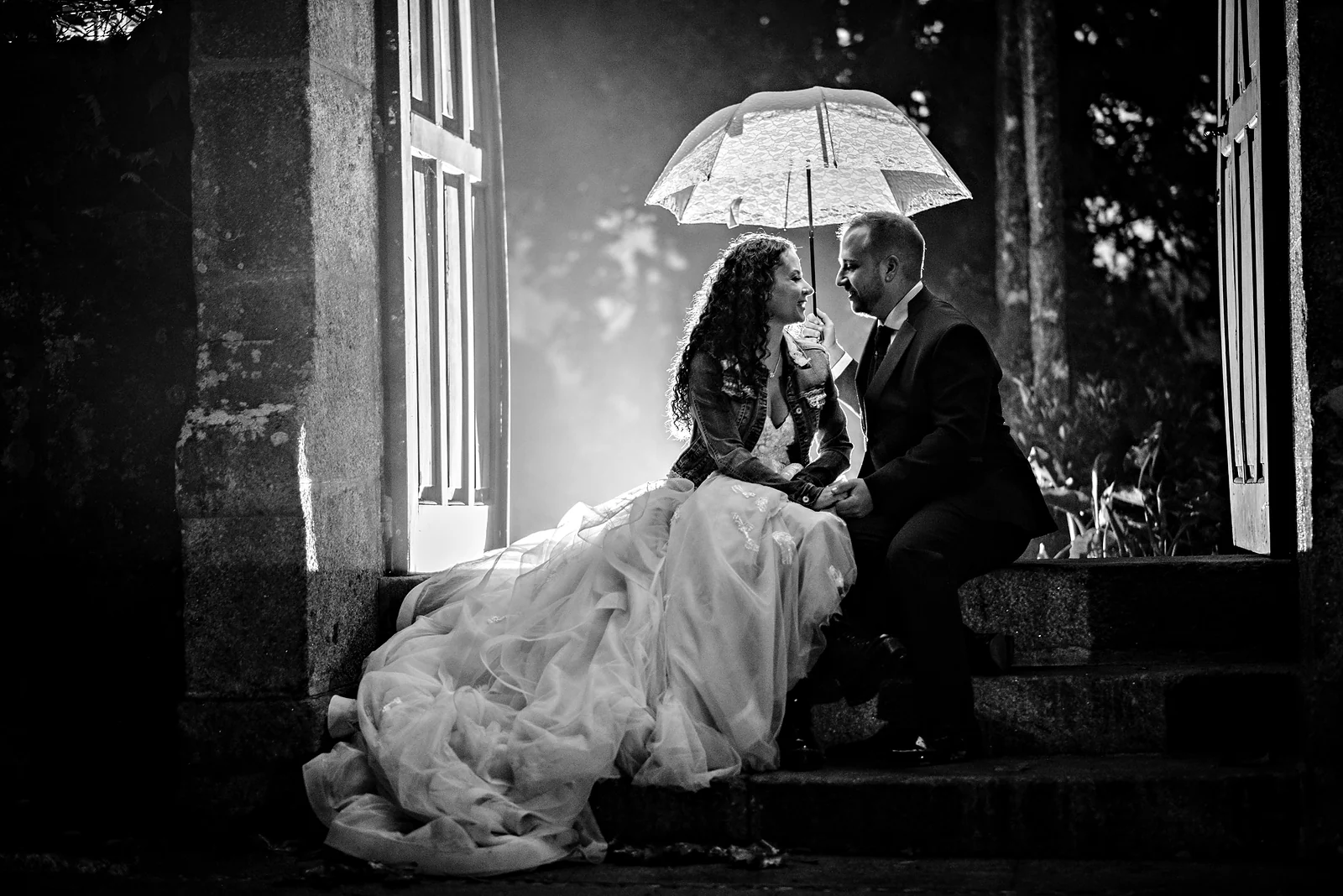 Pareja de novios bajo un paraguas, sentados en la escalera de piedra de un pazo, en blanco y negro.
