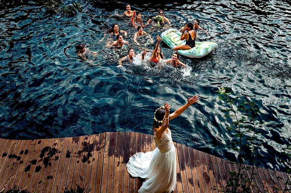 Novia de espaldas sobre una pasarela de madera lanzando el ramo a un grupo de invitadas que la esperan dentro del agua.
