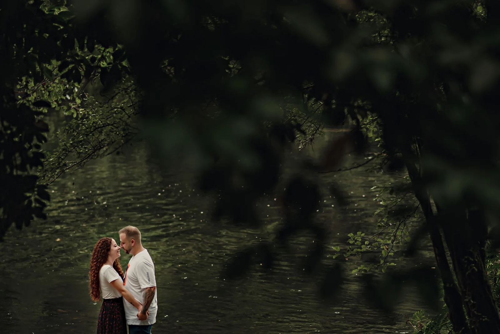 Pareja abrazada junto a un río, vista entre las ramas oscuras de un árbol en primer plano.