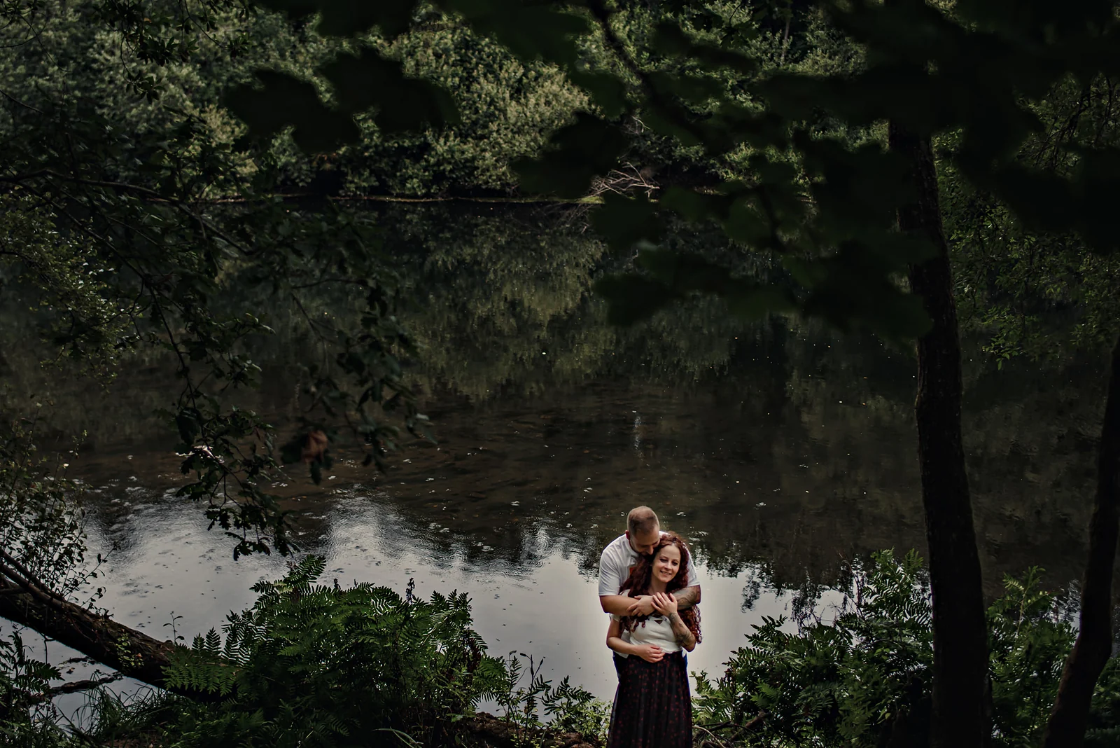Una pareja diminuta abrazada a la orilla de un río, rodeada de bosque y del reflejo del agua.